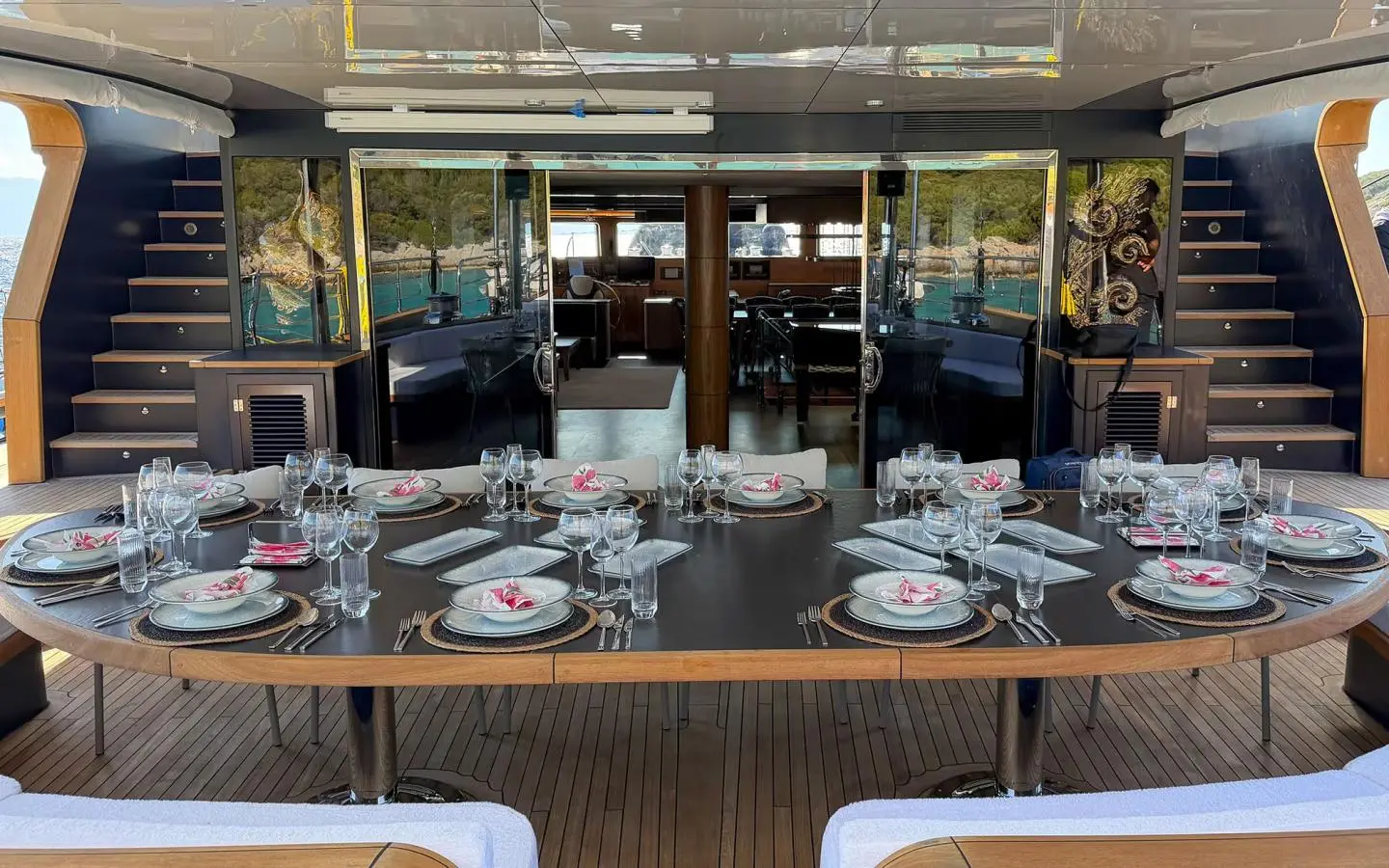 Large table set with plates and glassware on the yacht’s deck, with glass doors in the background.