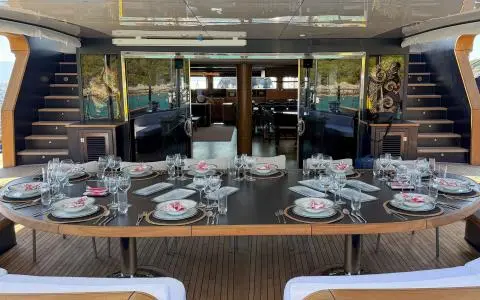 Large table set with plates and glassware on the yacht’s deck, with glass doors in the background.