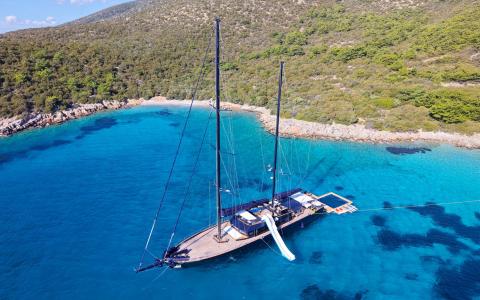 Sailing yacht with tall masts on turquoise blue water near a rocky green coastline