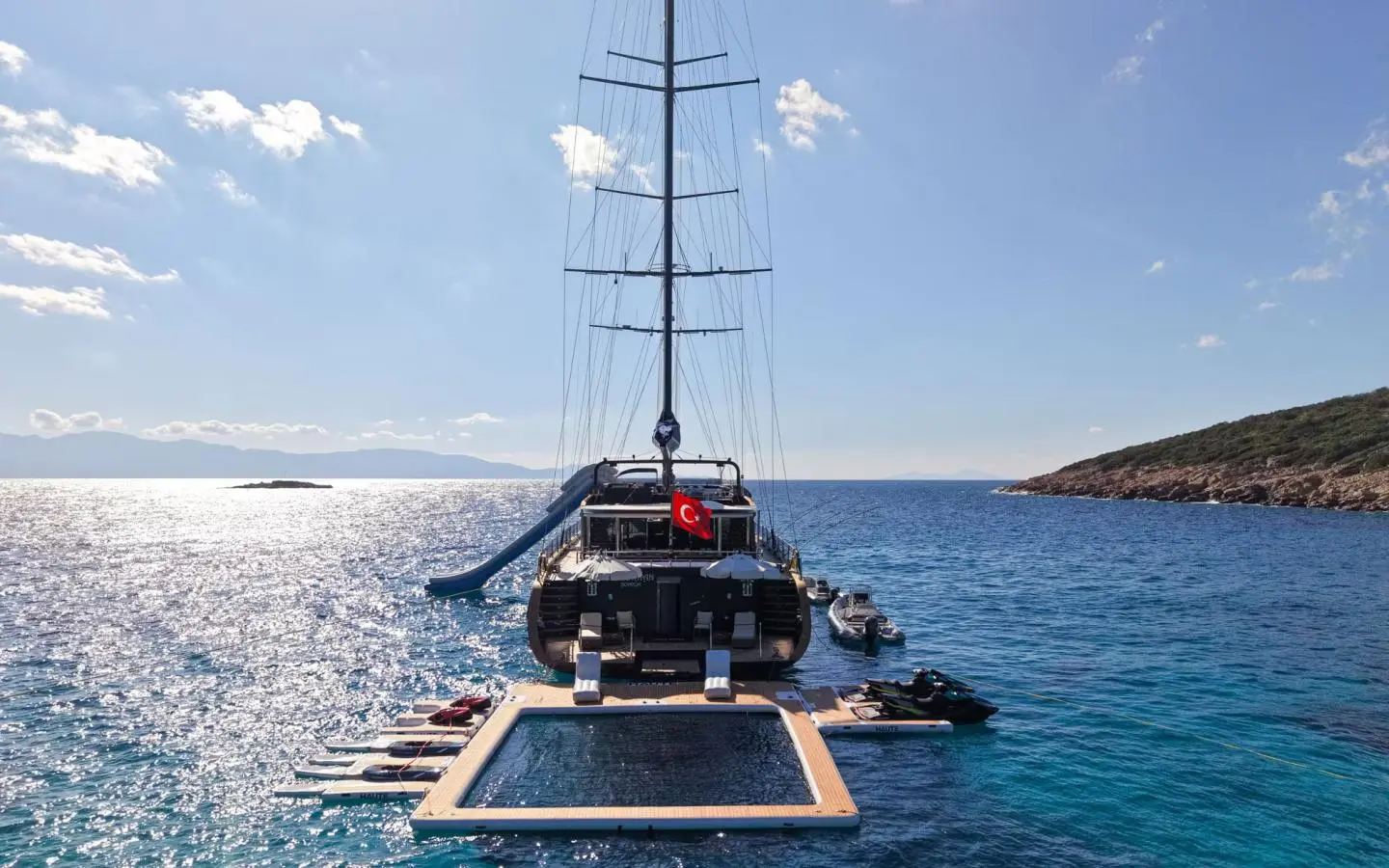 Sailing yacht with tall masts anchored in calm turquoise sea, slide on the side and a pool in the foreground