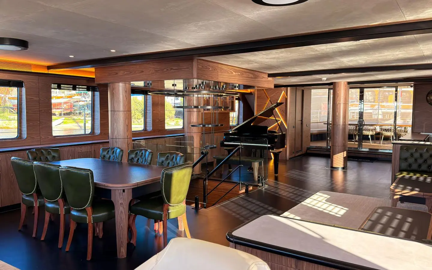 Indoor lounge with a large wooden table and green upholstered chairs, piano in the center, glass railing, and large windows