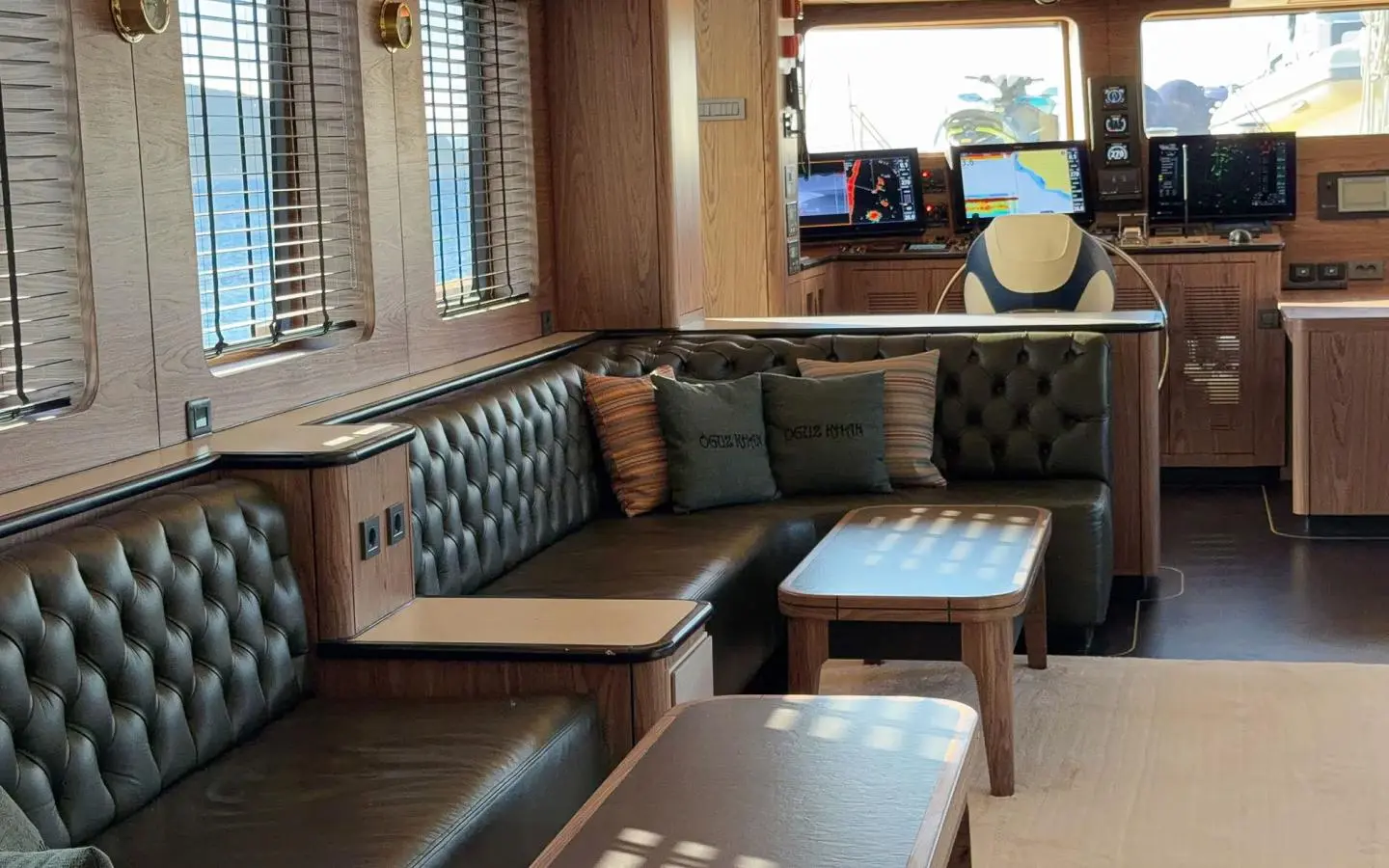 Indoor yacht lounge with black leather tufted seating, wooden panels, and white coffee tables, plus navigation screens in the background