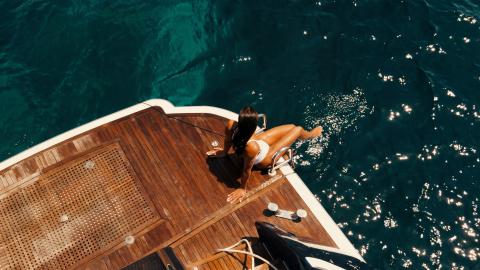 Woman in a white bikini sitting on a wooden yacht deck, dangling her legs over deep blue sea