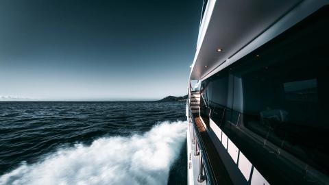 Right stern side of a luxury yacht cutting through water, sea spray and distant coastline visible