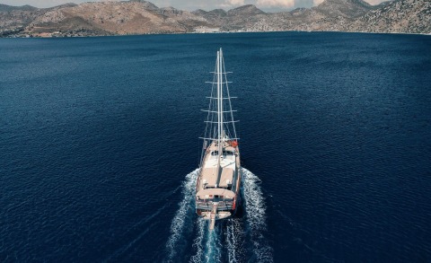 Gulet Nevra Queen with tall mast sailing in open sea, coastal hills in the background