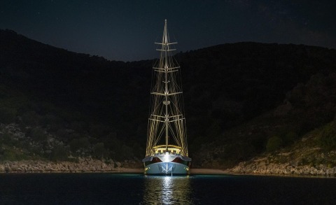Nighttime shot of a sleek sailing yacht moored in a dark cove with illuminated bow and tall masts