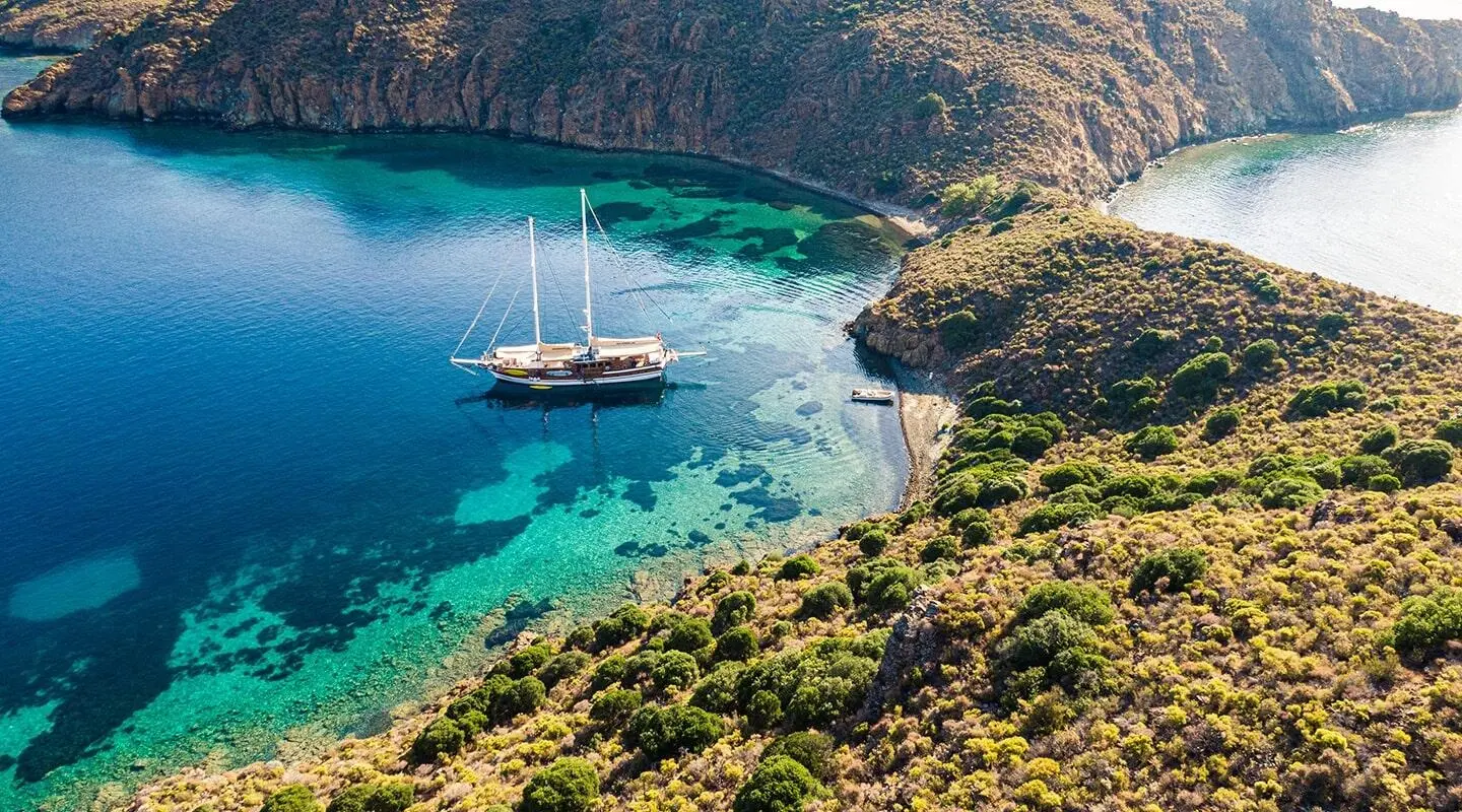 Derin Deniz yacht anchored along a rocky coastline at sunset with calm turquoise water