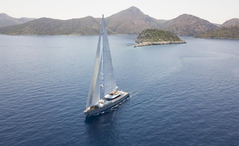 A two-masted sailing yacht sails through blue sea with rocky hills and a small island in the background