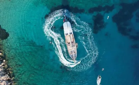 Aerial view of a blue and white gulet anchored in turquoise water near a rocky shoreline