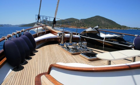 On the yacht's teak deck with chrome fittings, a black Yamaha jet ski on the right, purple fenders on the left, and coastal hills in the background