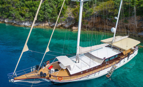 Luxury wooden-deck sailboat with tan canopy in turquoise water near rocky shore