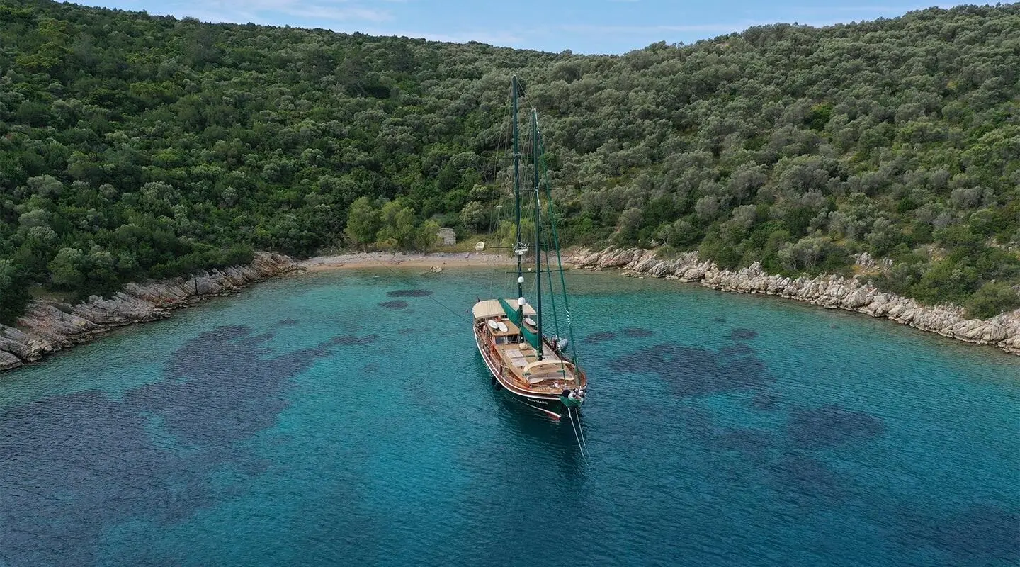 Ros Mare yacht anchored near a green hillside, calm blue sea at sunset