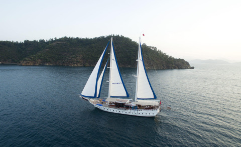 Two-masted sailing yacht with white hull and blue-trim sails near coastal hills