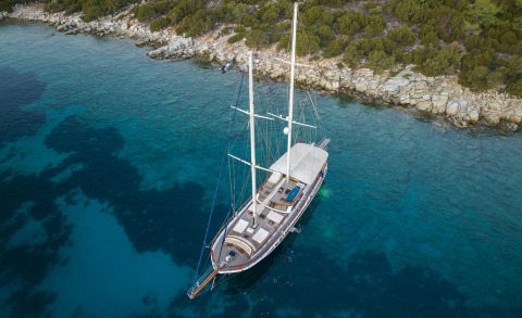 Two-masted sailing yacht afloat in turquoise water near rocky shore, with seating areas and a sunshade on the deck