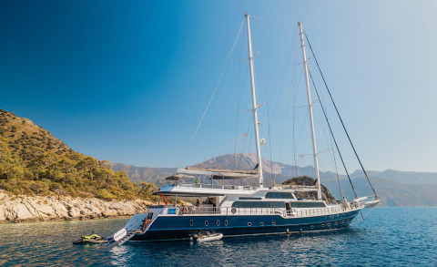 Blue motor-sail yacht anchored in clear sea, hull visible with two masts and rigging