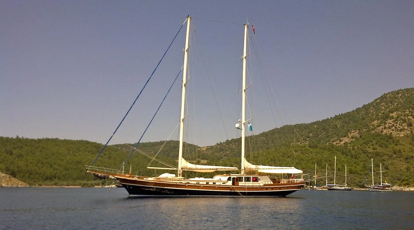 Two-masted wooden gulet yacht anchored on calm water by a hillside shore