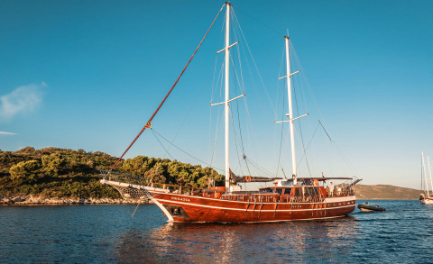 Gulet-style sailing yacht with two masts anchored near shore, people on the deck with calm sea