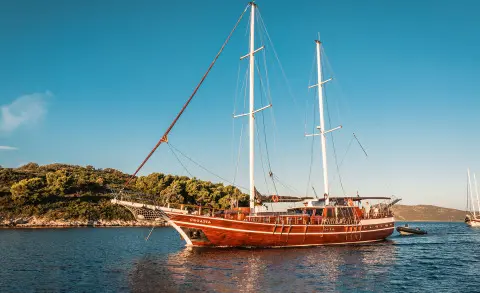 Yacht with tall masts anchored near rocky shore under clear blue sky