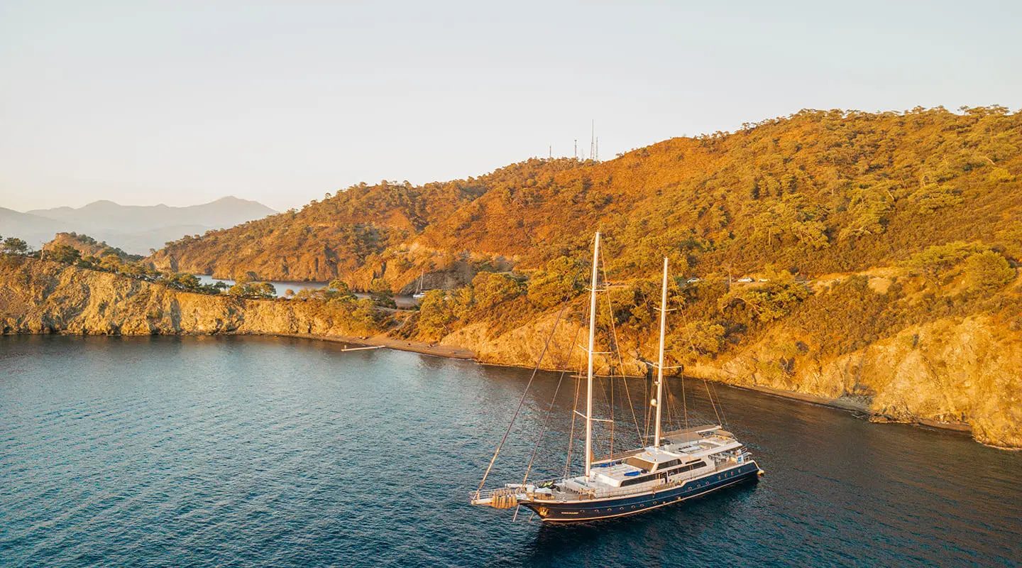 Gulet Esma Sultan anchored along coastline with rocky cliffs in sunset light