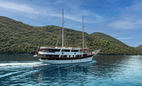 White wooden gulet yacht with two masts sailing on blue water, green hills in the background