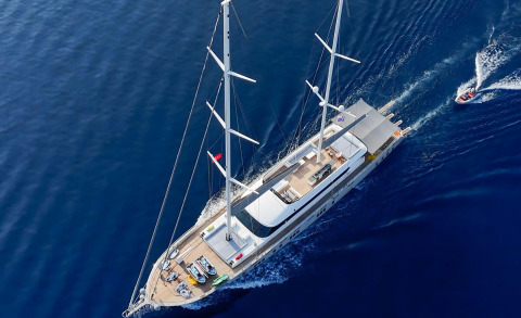 From above, a luxury two-masted sailing yacht with a visible deck, seating area, and equipment on the sundeck amid deep blue water.