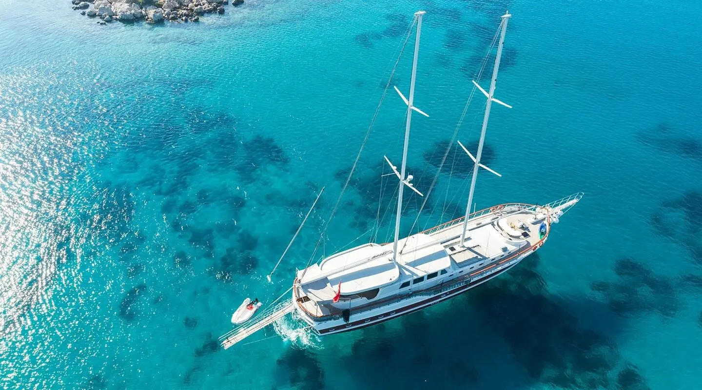 Aerial view of a white sailing yacht anchored in clear turquoise water near a rocky shoreline