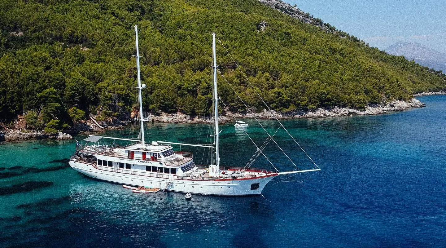 Two-masted yacht Corsario anchored near a green forested coast