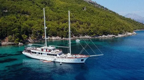Two-masted yacht Corsario anchored near a green forested coast