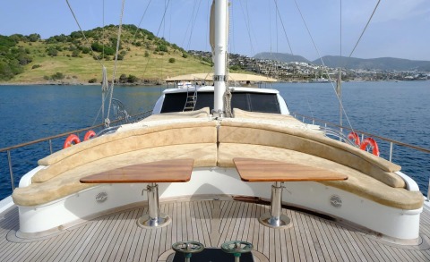 Forward view from the deck with blue sea, beige curved seating, two wooden tables, and mast cables against a shoreline with hills and buildings