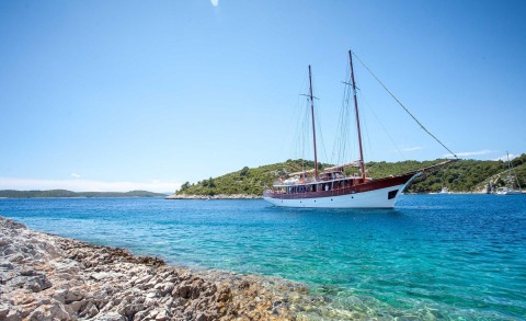 Two-masted sailing yacht with white hull anchored in clear blue water near rocky shoreline