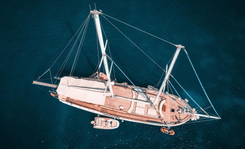 From above, Wicked Felina sails in clear blue water with a wooden deck and white rigging.