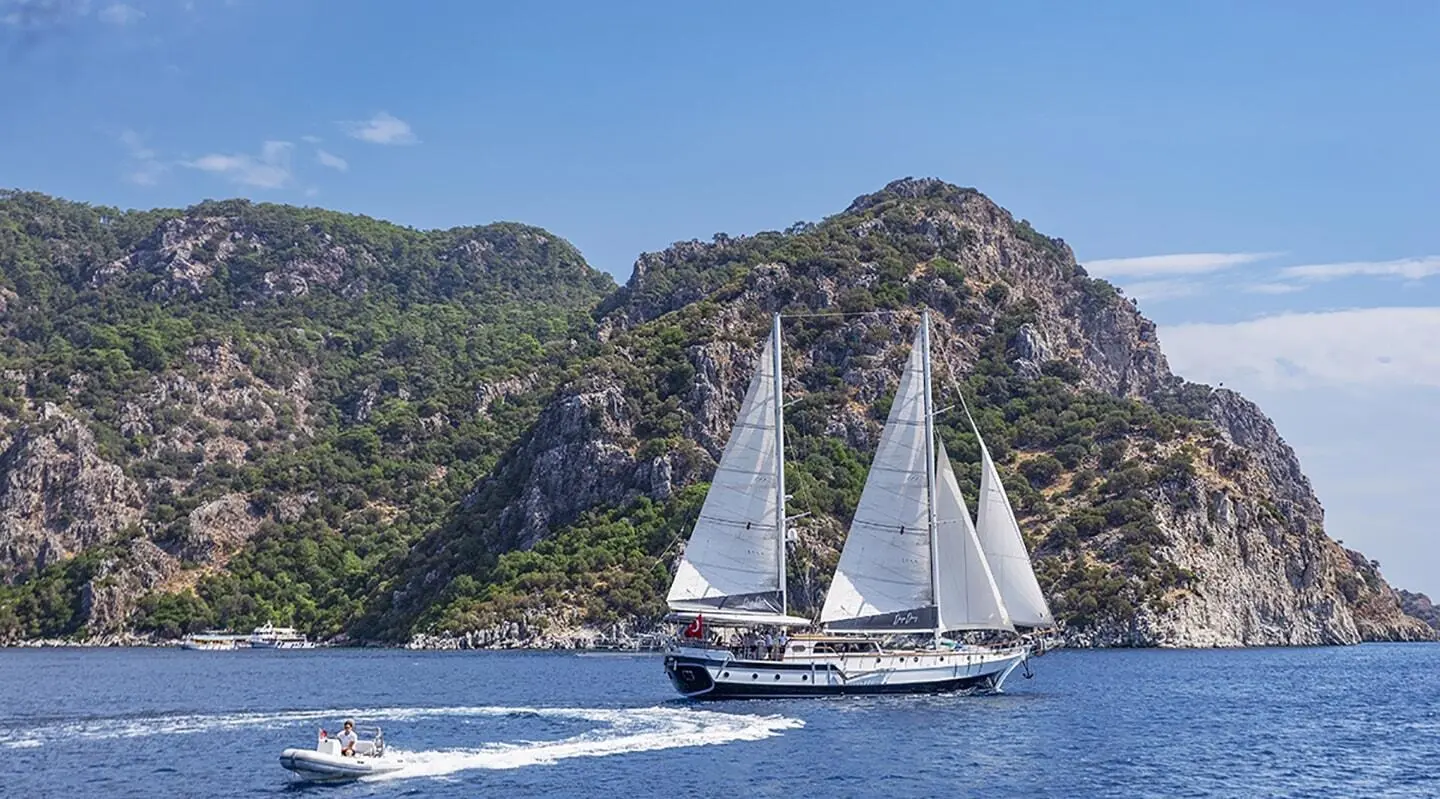 Yacht Derya Deniz sails near rocky cliffs at sunset with two white sails