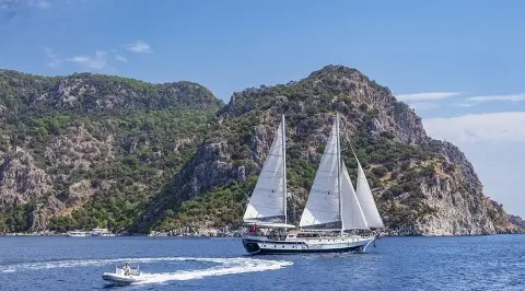 Yacht Derya Deniz sails near rocky cliffs at sunset with two white sails