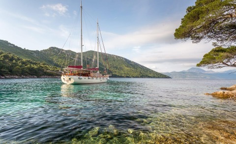 Yellow-white sailing yacht anchored in clear blue water near rocky shoreline