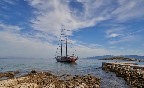 Wooden sailing yacht anchored near rocky shore under a blue sky