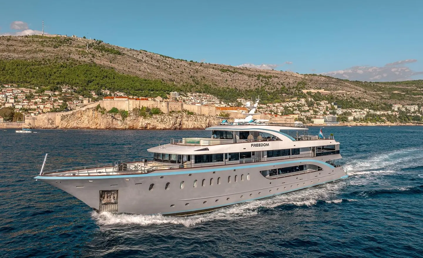 Luxurious gray yacht Freedom sailing near rocky coastline under blue sky