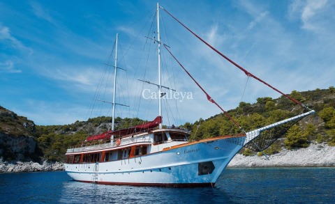 Aerial view of Cataleya tall-masted motor-sail yacht anchored in clear blue sea, with sunlit deck laid out for luxury cruising