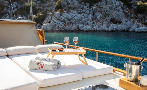 Deck of a luxury yacht with two wine glasses and snacks on a small table, blue sea and rocky coastline in the background