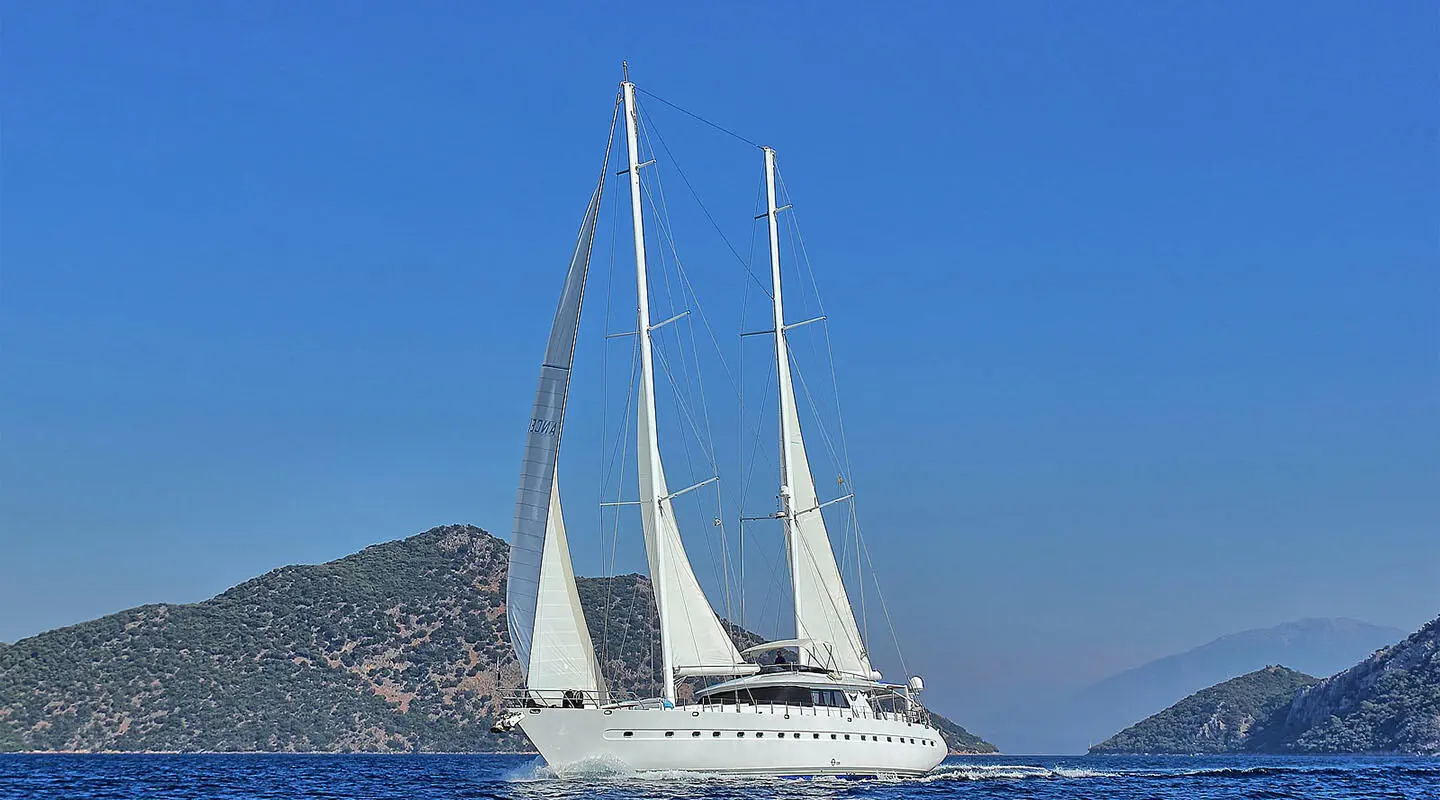 Two-masted white sailing yacht on open sea, with mountainous coast in the background