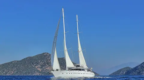 Two-masted white sailing yacht on open sea, with mountainous coast in the background