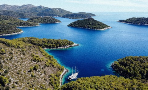 A three-masted sailing yacht anchored in a blue bay surrounded by green hills and clear water
