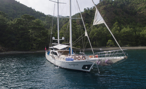 White sailing yacht anchored near forested rocky shore, with deck life rings and rigging visible