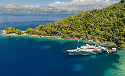 Maia yacht anchored in calm blue waters near a green forested coastline with mountains in the background