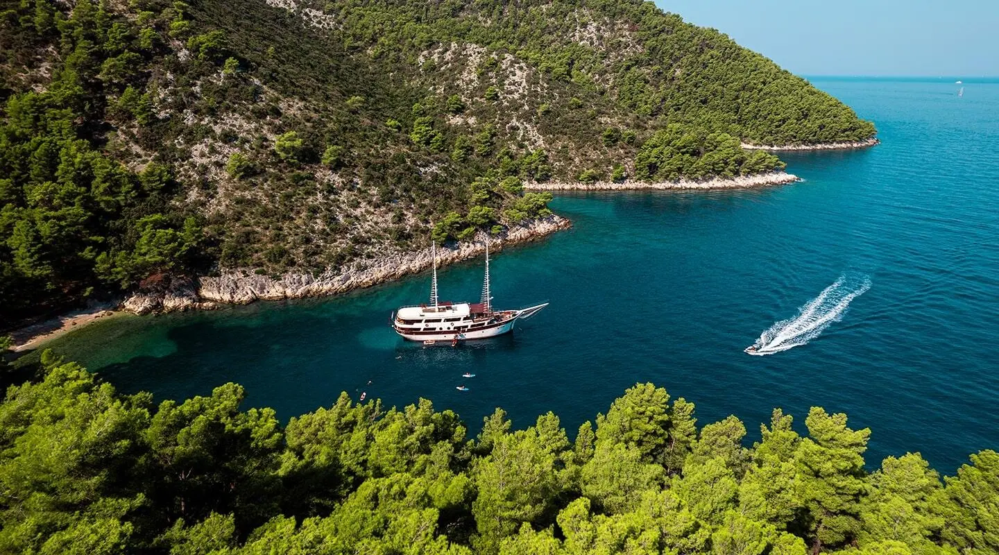 White double-masted gulet yacht anchored in a calm cove with green hills