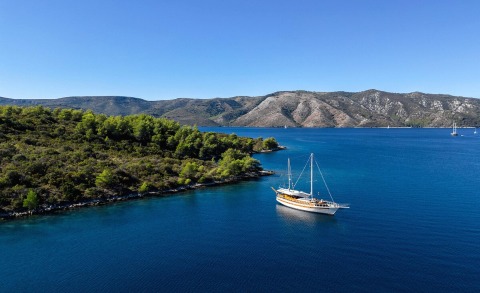 A gulet sailboat anchored in blue calm water near a green forested coastline