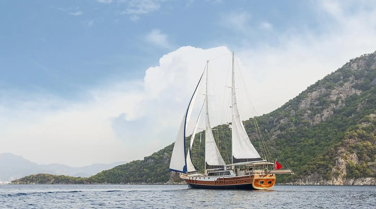 Diva Deniz yacht with white sails near a rocky coastline at sunset