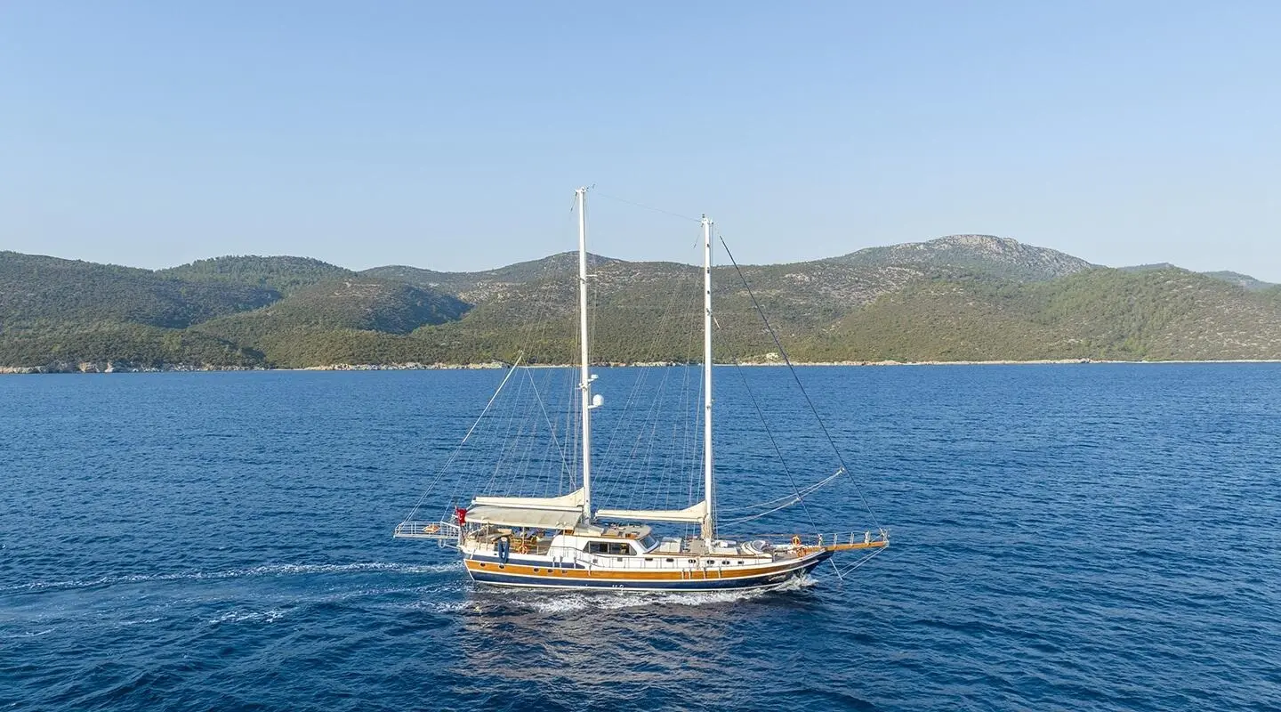 Two-masted luxury sailing yacht on blue sea with a coastal hillside in the background