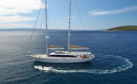 Acapella sailing yacht anchored offshore with a tender cruising beside, azure sea and distant coastline