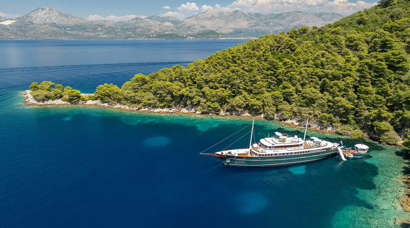 Maia yacht anchored in a small turquoise bay beside a green forested coastline
