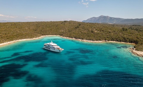 Olimp yacht anchored in turquoise waters near a sandy bay with rocky shoreline